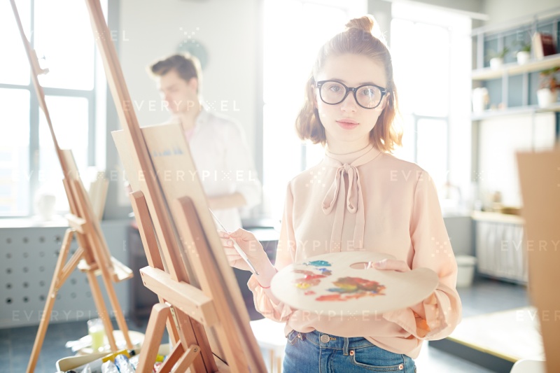 Pretty girl in eyeglasses and smart casual looking at camera at lesson of painting in modern school of arts